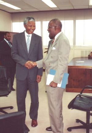In an office setting, two men stand smiling at each other and shaking hands. The man on the right is wearing a grey suit and holds a dossier of papers. The man on the left is Nelson Mandela.