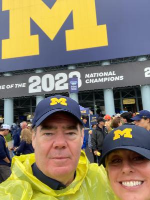 Two people stand next to each other smiling in a football stadium. They both wear navy blue baseball hats with a block M in the middle. Behind them, a sign appears with a large block M and below it, a banner that reads “2023 National Champions.”