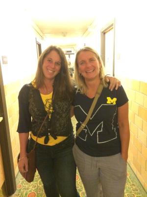 Two women stand with their arms around each other in a hallway. They are wearing University of Michigan maize and blue apparel.