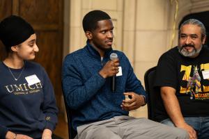 One Detroit community member sits between two people and speaks into a microphone 