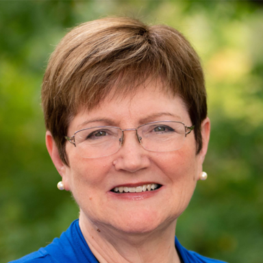 Headshot of adult wearing bright blue blazer and glasses, smiling, outdoors.