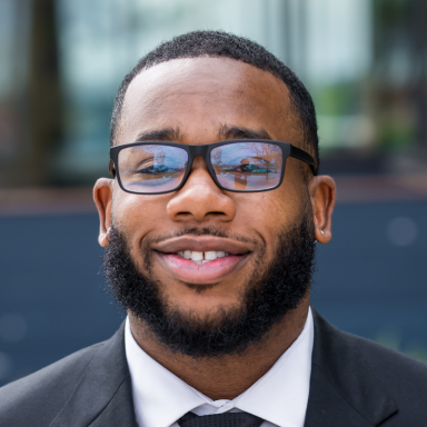 Headshot of adult wearing black suit jacket and tie and glasses, smiling, outdoors.