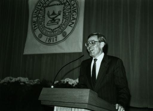 Black and white photo of Cecil Miskel, wearing a suit and tie, standing at a podium during a graduation, laughing into the microphone.