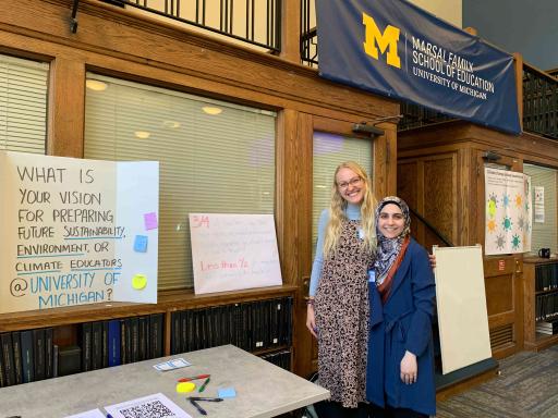 Two students stand smiling with their arms around each other in front of a display table. Behind them is a poster that reads “What is your vision for preparing future sustainability, environment, or climate educators @ University of Michigan?” 