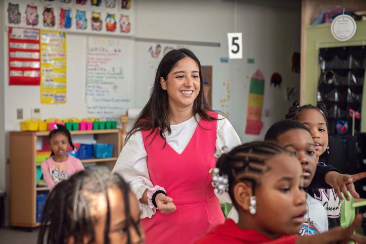 A teaching resident wearing a pink jumpsuit stands in a colorful classroom. Early elementary students are standing around her and reaching for something out of the photo frame.