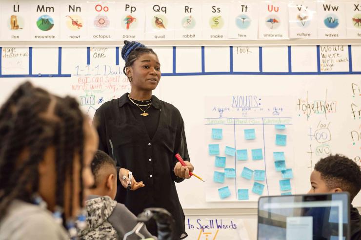 A teaching resident wearing a black blouse stands in front of a white board while three elementary school students listen to her.