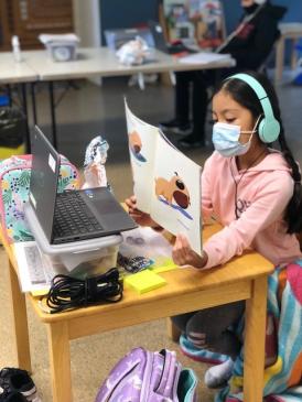 Girl reading to screen (Michigan Education Magazine, Fall 2020) Young person in pink sweatshirt wearing headphones over her ears and a surgical mask over her mouth shows the pages of a picture book to a laptop webcam