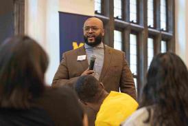 David Humphrey, dressed in a brown suit with houndstooth-patterned undershirt and holding a microphone, speaks to a group of students