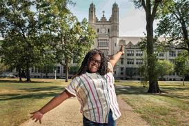 Teenager, wearing a white shirt with vertical stripes in primary colors and jeans, with long locs, shown from the waste up, with their arms outstretched, smiling at the camera. The Marygrove building's entrance towers in the background. 