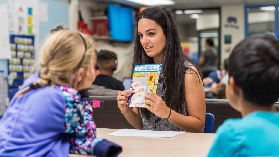 A young woman with long brown hair sits across from two elementary school students, pointing to details on the cover of a book. They are situated in a classroom with children at other desks behind them.