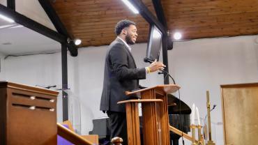 Eric Veal Jr. wears a black suit and speaks from behind a podium on a stage.