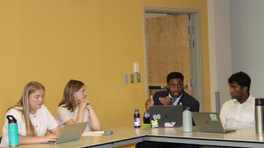 Eric Veal Jr. gestures with his hands as he speaks with fellow students in a meeting setting. The students sit at two tables that have been pushed together and type on laptops.