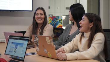 Victoria Vezaldenos sits at a desk with students. They are all typing on laptops, and looking up from their work to laugh together.