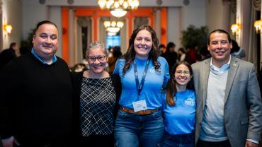 Victoria Vezaldenos poses in a blue t-shirt that reads 'Puentes." She stands in the middle of five people dressed in business attire who have their arms around each other, smiling at the camera.