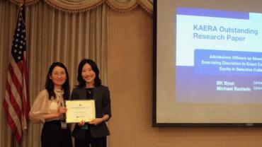BK Byun is pictured on stage accepting a certificate for the KAERA Outstanding Research Paper award. Wearing professional attire, she stands next to a person administering the award. Behind them and to the left an American flag stands in a flagpole; behind them and to the right, a screen announces the name of the award.