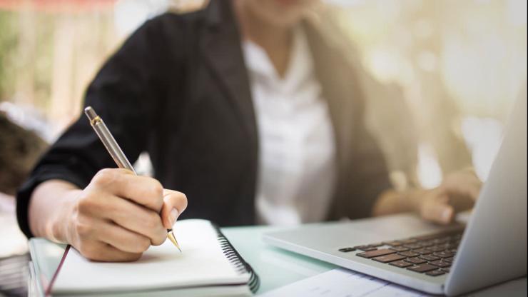 person writing notes on a note pad with a pen and using a laptop with their other hand