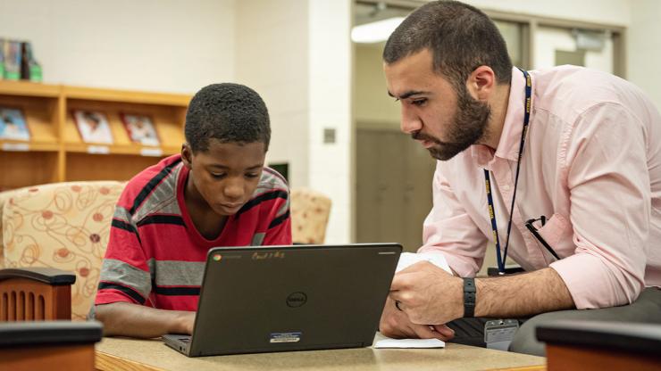 A male teacher helps a male student at a laptop