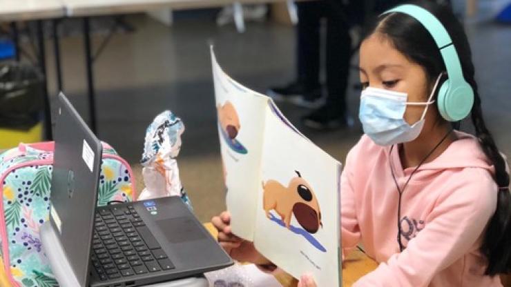  Young person in pink sweatshirt wearing headphones over her ears and a surgical mask over her mouth shows the pages of a picture book to a laptop webcam
