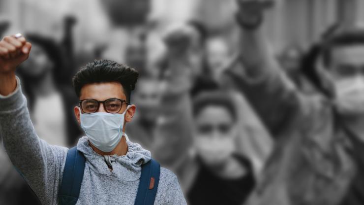 A black and white photo of a group of protesters wearing surgical masks and raising their fists. One protester stands in the foreground, in color.