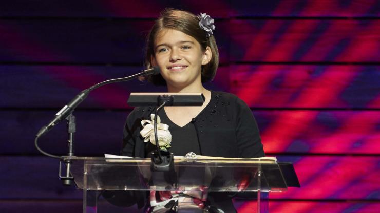 Photo of Olive Martin. Olive wears a dress with a black top and floral bottom, and smiles while standing in front of a podium.