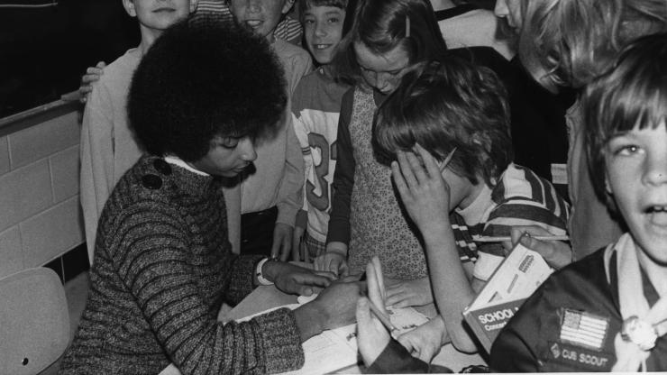 Black and white photo of Rita Woods helping a student with a workbook while a group of students congregates around her