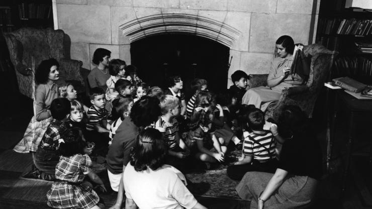 Black and white photo of University Elementary School students sitting on the floor in front of a fireplace as a teacher reads to them, 1949. 