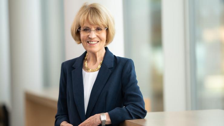 Mary Sue Coleman, wearing glasses, a white blouse, a navy blue jacket, and gold necklace, smiles and poses indoors, with one arm on a banister and her hands clasped in front of her, for a photograph.