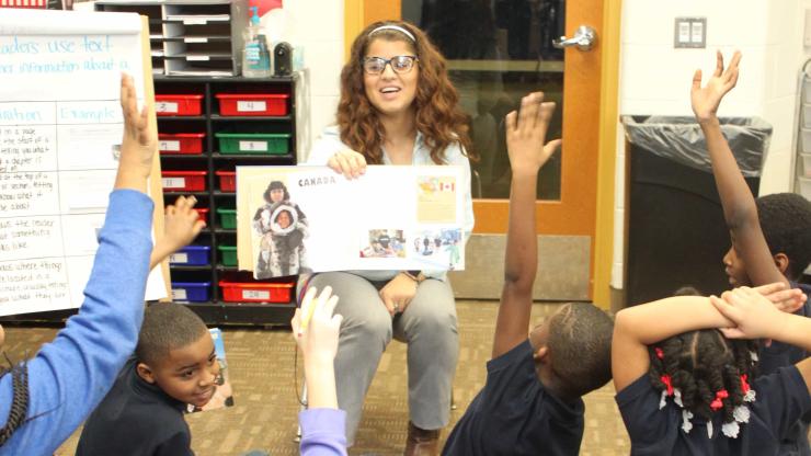 Student teacher happily shows pages of a book to group of children excitedly raising their hands.