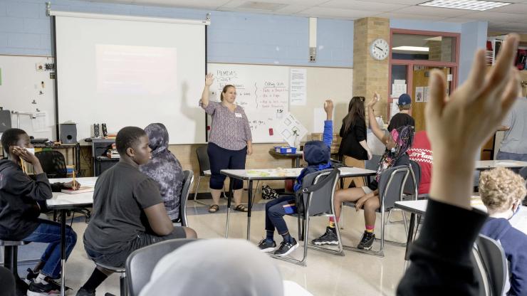 Teacher raising her hand stands at the front of a classroom full of students