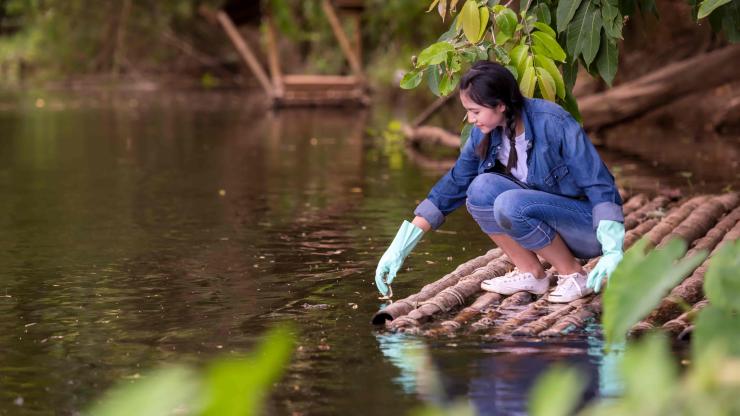 A student on a raft made of logs, wearing gloves, reaches into water.