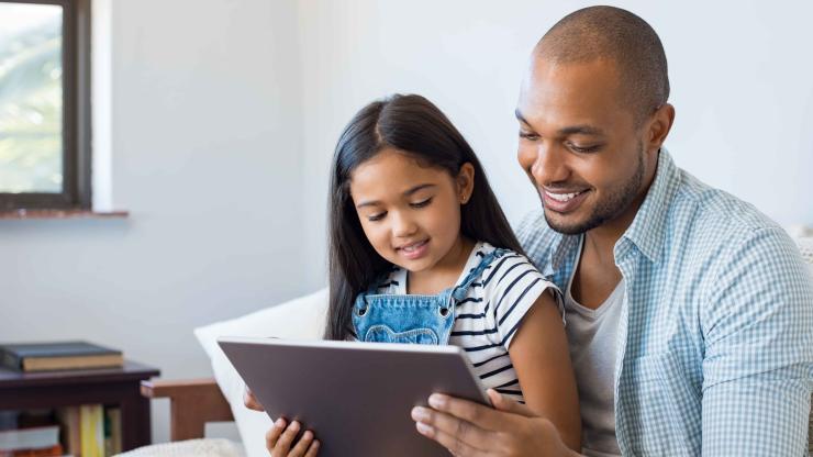 Father and smiling daughter sitting on sofa using digital tablet.