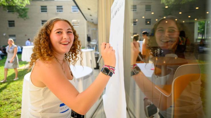 A student in a courtyard writes on a large sheet of paper taped to an exterior window.