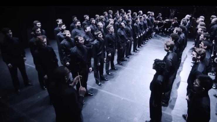 A large choir, divided into several single-file lines facing each other, all dressed in black attire. A conductor, also in black, leads the group with his baton raised. In the background, an orchestra waits to perform. Bright white lighting focuses on the choir and creates stark shadows.