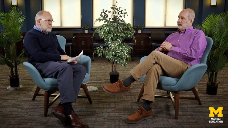 Don Peurach and Barry Fishman sit across from each other in blue armchairs, having a conversation in a formal setting with plants and large windows in the background.
