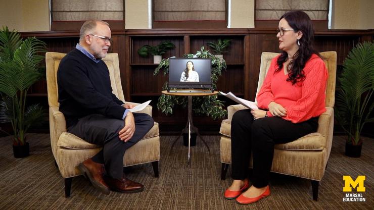 Don Peurach and Rebecca Quintana sit in beige chairs facing each other, with plants surrounding the scene. Between them is a small round table with a video monitor showing an AI-generated avatar of Rebecca.