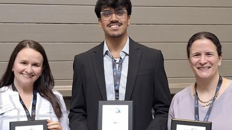 Three people stand in a row, displaying plaques that announce awards. Marsal School ELMAC student Ongkon Deb stands in the middle in a black blazer, wearing a lanyard and glasses.