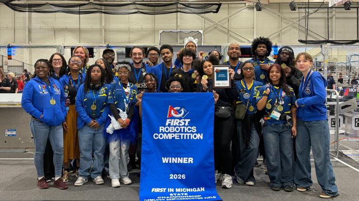 A large group of high school students poses together in a warehouse. The students wear blue t-shirts and hoodies, with medals around their necks. The student in the front center position holds a vertical blue banner that reads "FIRST Robotics Competition: Winner 2026."