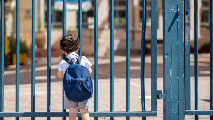 An elementary school child wearing a white t-shirt, grey shorts, and a blue backpack stands facing a closed gate. In the distance behind the gate a closed school with a brick facade is visitbe.