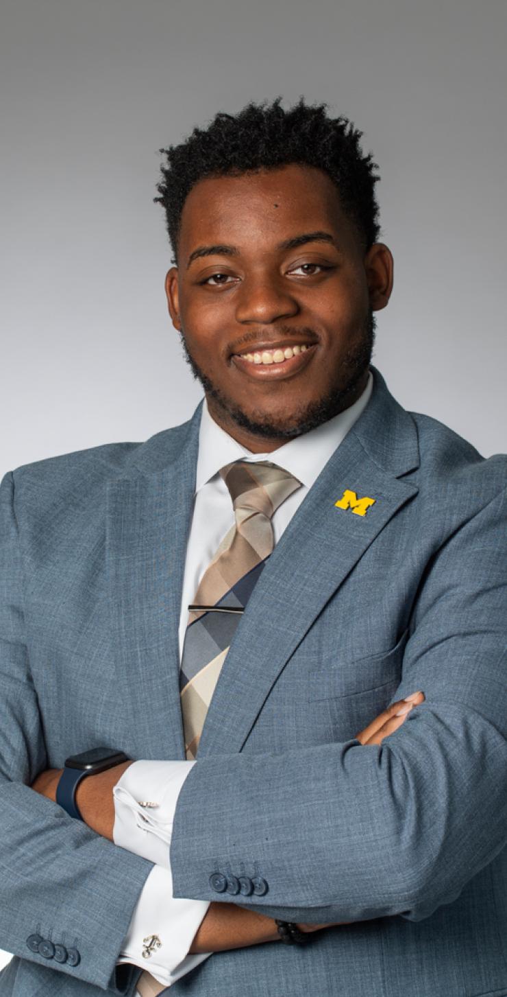 Headshot of Eric Veal Jr., who stands before a grey background in a grey-blue blazer with a block M pin pinned to his lapel.