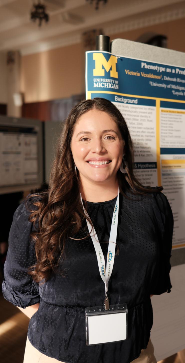 Victoria Vezaldenos stands before a poster at a conference. She wears a black blouse with a white lanyard around her neck.