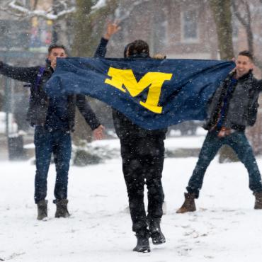 Four students having fun on a snowy day in Ann Arbor. One student carries a small flag with the block M on it.