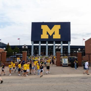 An enormous scoreboard in the background with the maize block M on a blue background. Students wearing maize and blue file out from an exit beneath the scoreboard.