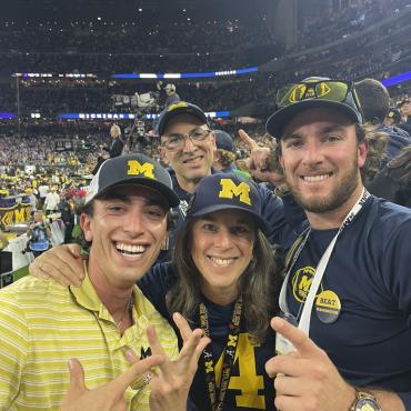 A woman stands in a stadium with her two young adult sons on either side of her, and her husband just behind her. They all wear University of Michigan maize and blue apparel. 