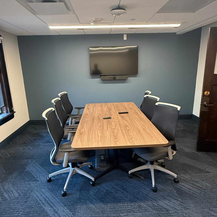 Image of conference room, with one table surrounded by chairs, and a monitor on the far wall