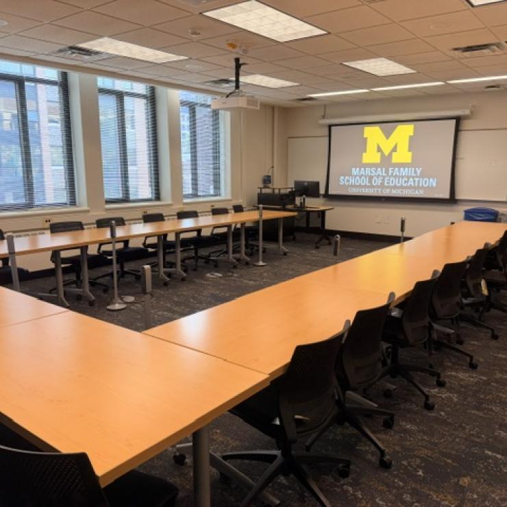 A classroom with desks and chairs with a projector screen with a block M. 