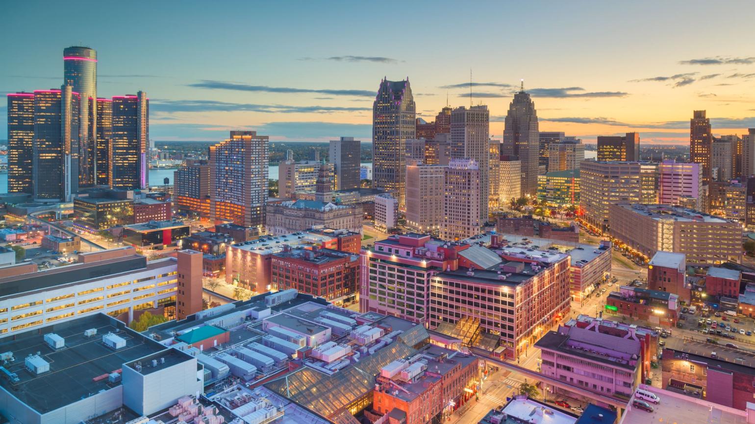 Detroit Skyline at sunset. On the left side of the skyline, the Renaissance center's towers glow pink. Center right, the Ally Detroit Center and Fischer Building rise above the Detroit River. In the foreground, smaller office buildings and parking garages glow pink from the sunset.