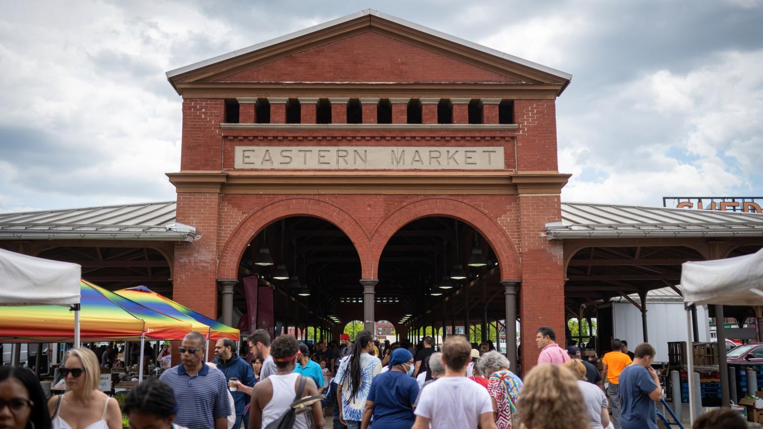 A summer crowd moves beneath a brick pavilion that rises in the background. The pavilion has a sign that says Eastern Market.