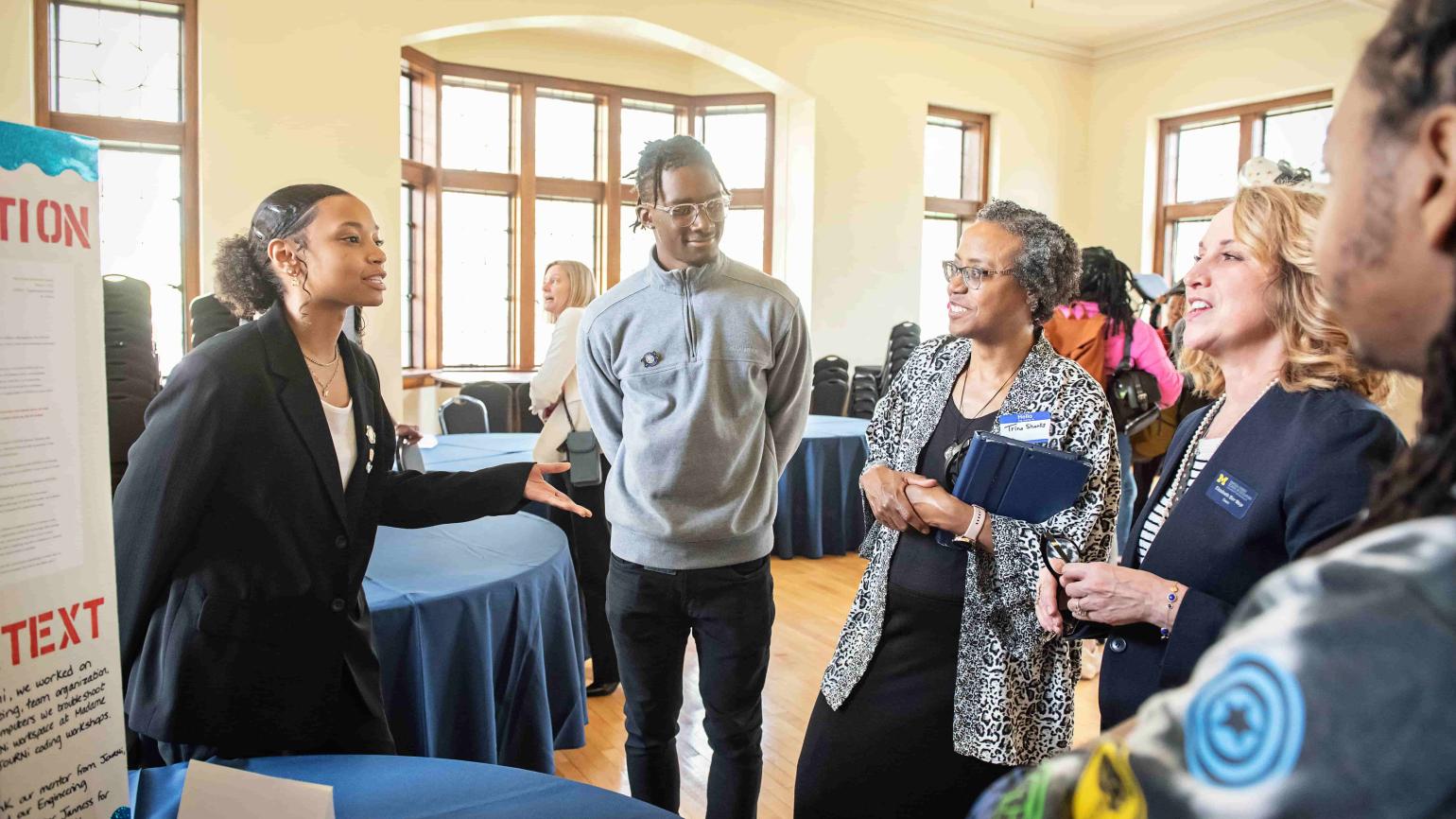 Two students stand beside a poster board in a bright room with large windows. One student, wearing glasses, a grey jumper, and black slacks, stands with his hands behind his back and smiles at three onlookers who listen to the other student, who wears a black blazer with a white blouse and speaks while gesturing with her hands.