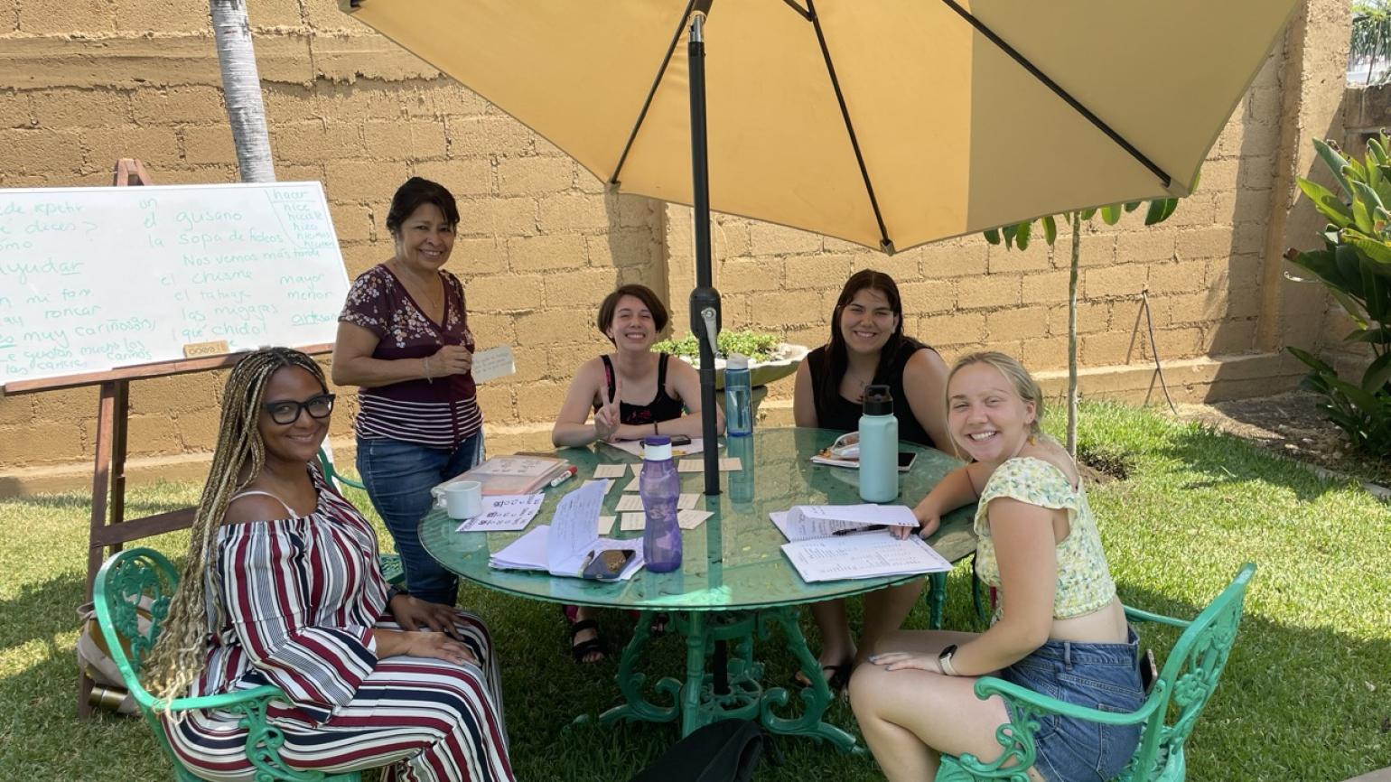 Teaching interns gathered around a table under a large umbrella for an outdoor Spanish lesson
