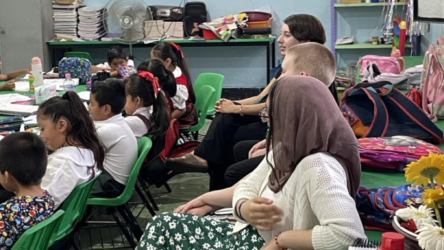 Three interns sit in the back of a Mexican school classroom and watch the teacher deliver a lesson to a group of students, seated in the row in front of them.